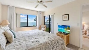 A bedroom with a bed, a ceiling fan, a TV on a wooden stand, and a view of the sea through a window.