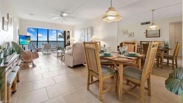 A spacious living area with a dining table set for four, surrounded by wooden chairs. A ceiling fan and pendant lights are visible. The room has a large window offering a view of the outdoors.