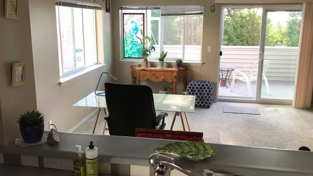 Ein Zimmer mit Glastisch, schwarzem Sessel, Nachttisch aus Holz mit Pflanzen und Blick auf eine Terrasse mit Stuhl und Tisch.