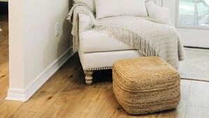 Un fauteuil blanc agrémenté d’un plaid et d’un pouf en osier, dans une pièce au plancher en bois et à la porte blanche.