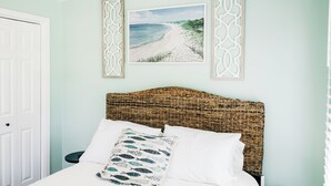 Une chambre à coucher avec un lit, des oreillers blancs, une tête de lit en osier, un tableau représentant une plage, encadré, et une porte blanche.