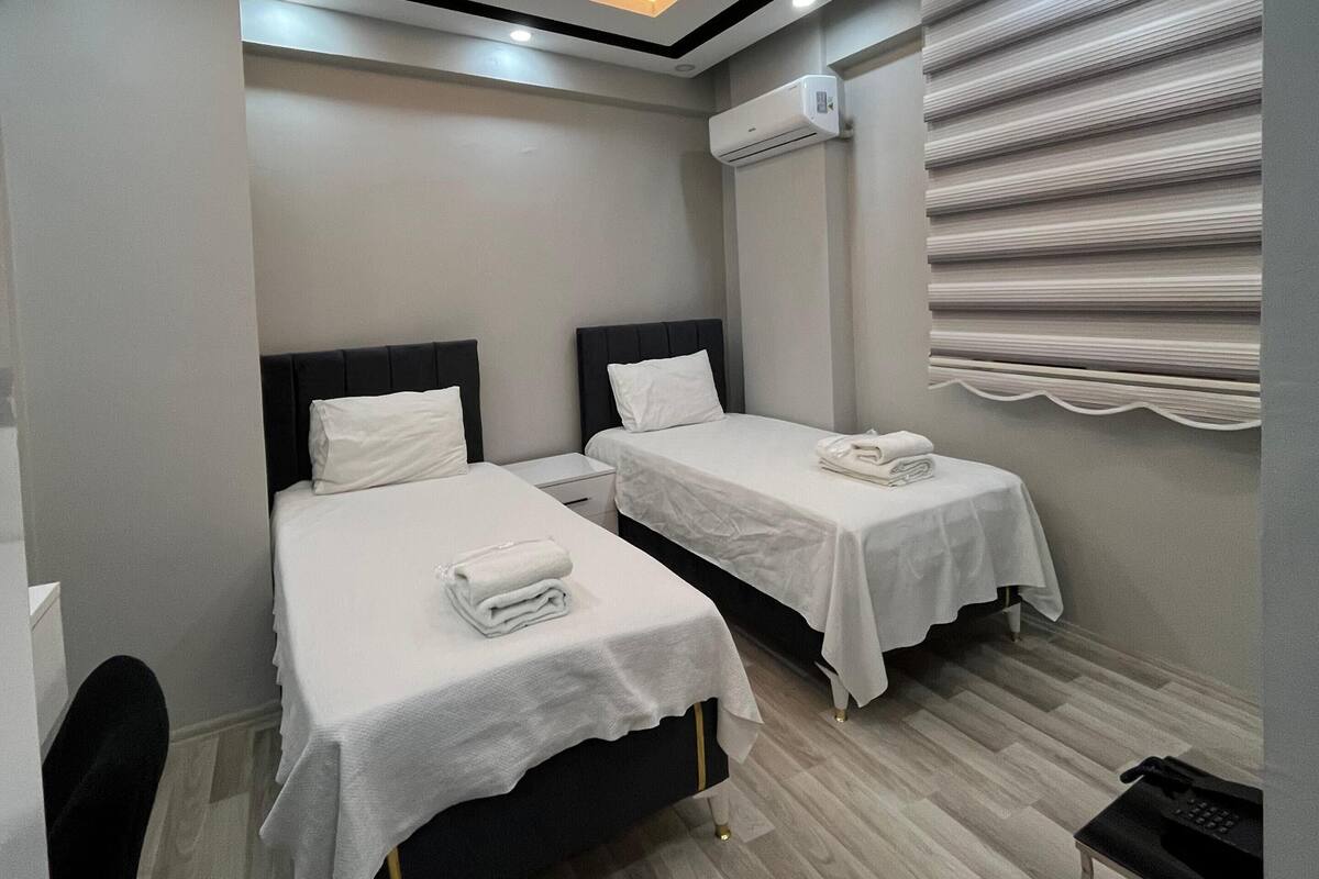 A hotel room with two beds, a desk, a chair, an air conditioning unit, and a window with blinds.