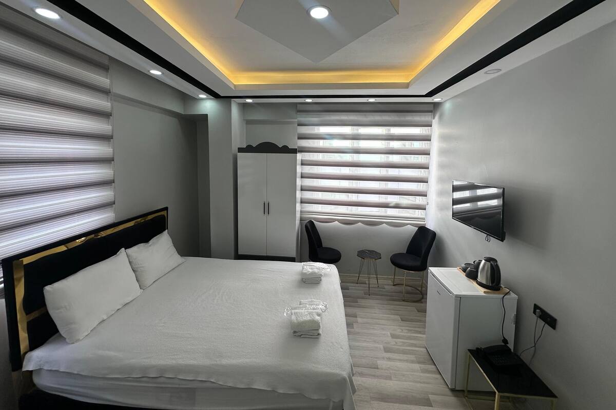 A modern hotel room with a neatly made bed, a desk with a kettle, a TV mounted on the wall, and a wardrobe.