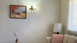 Een woonkamer met een glazen tafel, een beige bank, een lamp, een schilderij en een raam met jaloezieën.