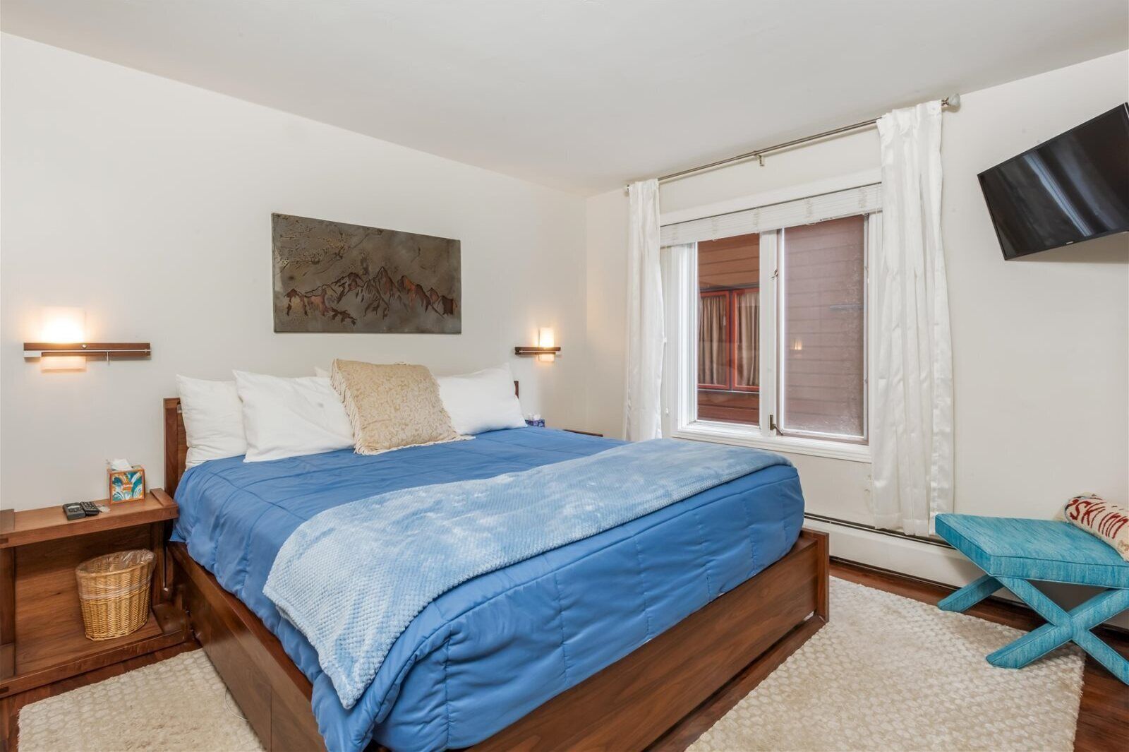 A bedroom with a wooden bed, a painting on the wall, a TV mounted above the bed, and a window with curtains.
