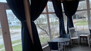 Een overdekt terras met een tafel en stoelen, grote ramen met gordijnen en lichtslingers.