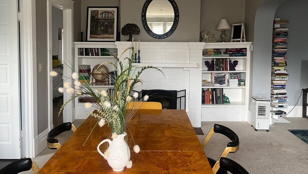 Een eetruimte met een houten tafel, stoelen, een witte kruik met bloemen, een open haard, boekenplanken en een spiegel aan de muur.