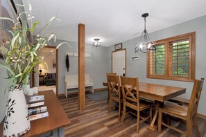 A dining area with a wooden table and chairs, a vase with greenery, and a window with blinds. - Aspen 2 | Fireplace, Restaurant, Pool, Spa (Blowing Rock)
