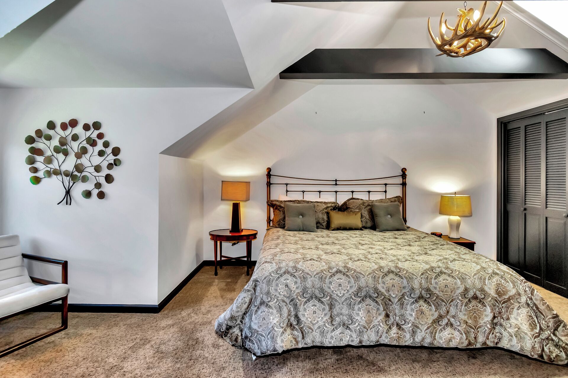 Um quarto com teto inclinado, uma cama com roupa de cama estampada, duas luminárias de cabeceira, um ornamento de árvore fixado na parede e um lustre.