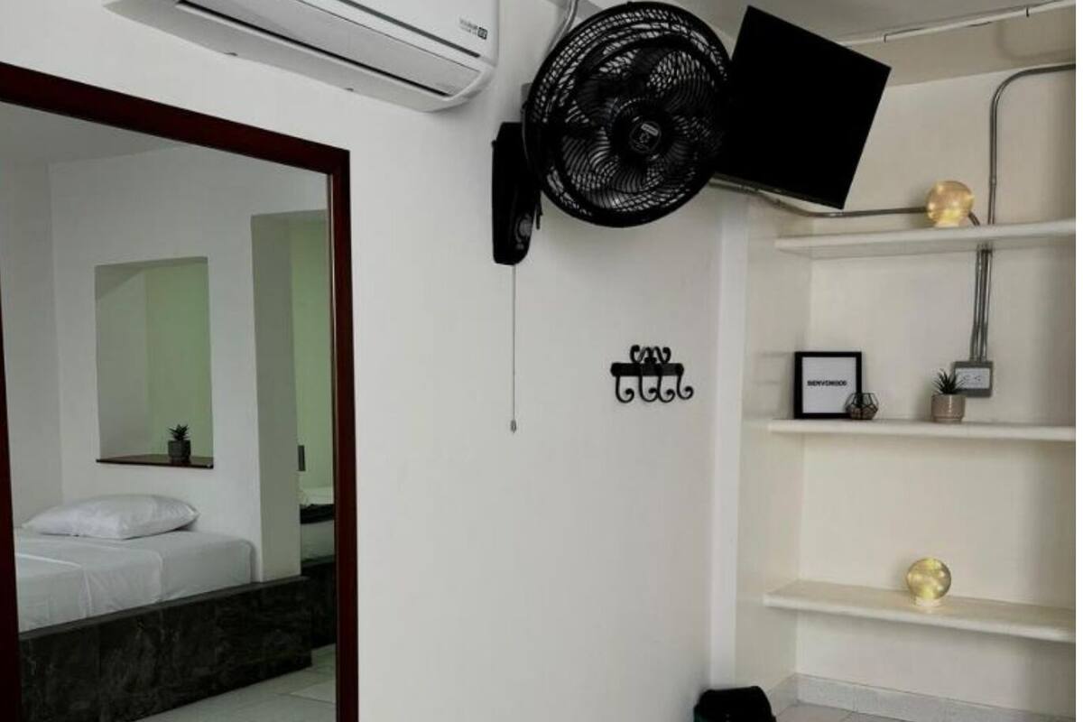 Une chambre d’hôtel équipée d’un lit, d’un climatiseur fixé au mur, d’un ventilateur sur pied, d’un miroir et d’une petite poubelle.