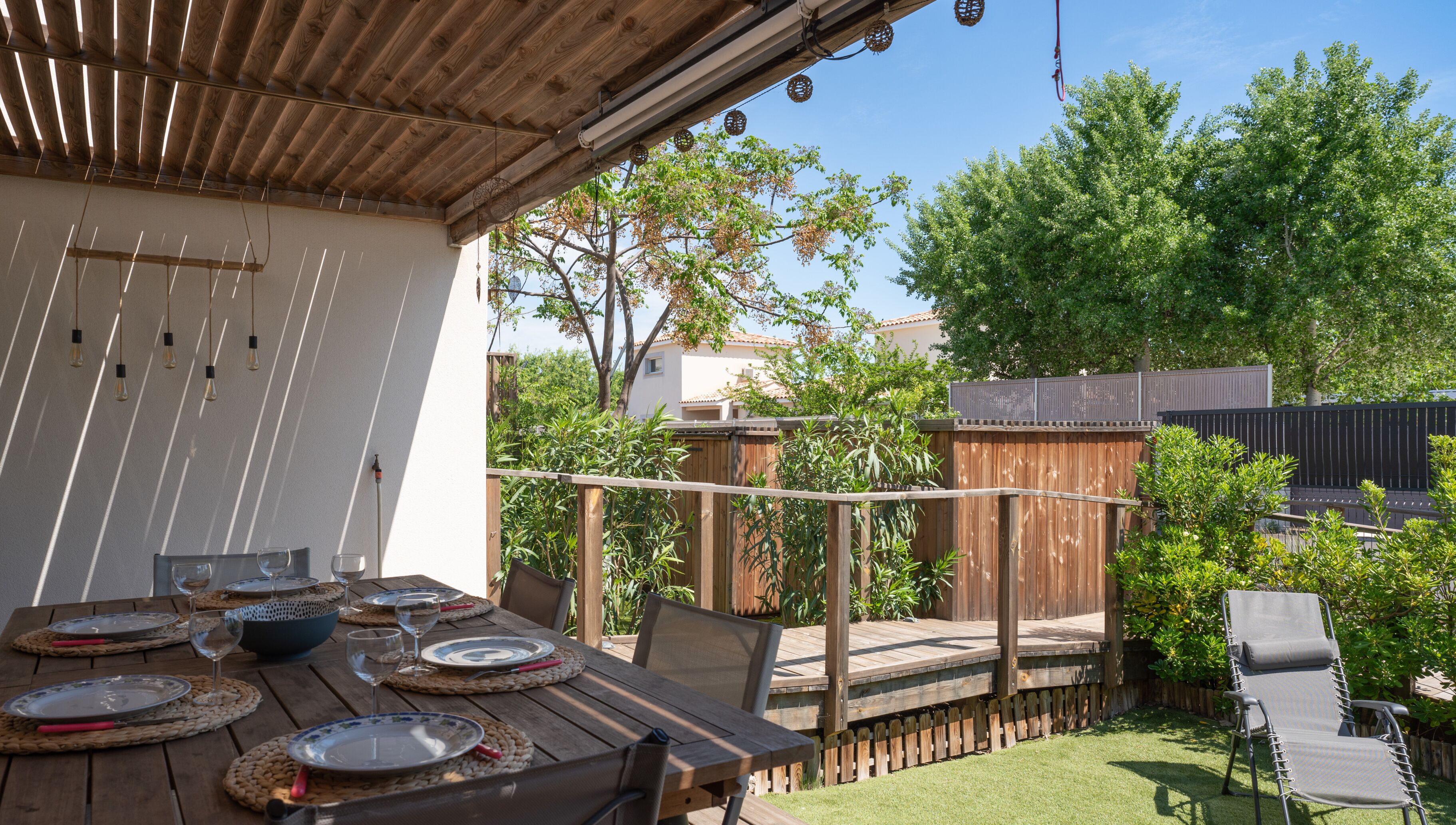 A wooden deck with a table set for dining, surrounded by a wooden fence and greenery.