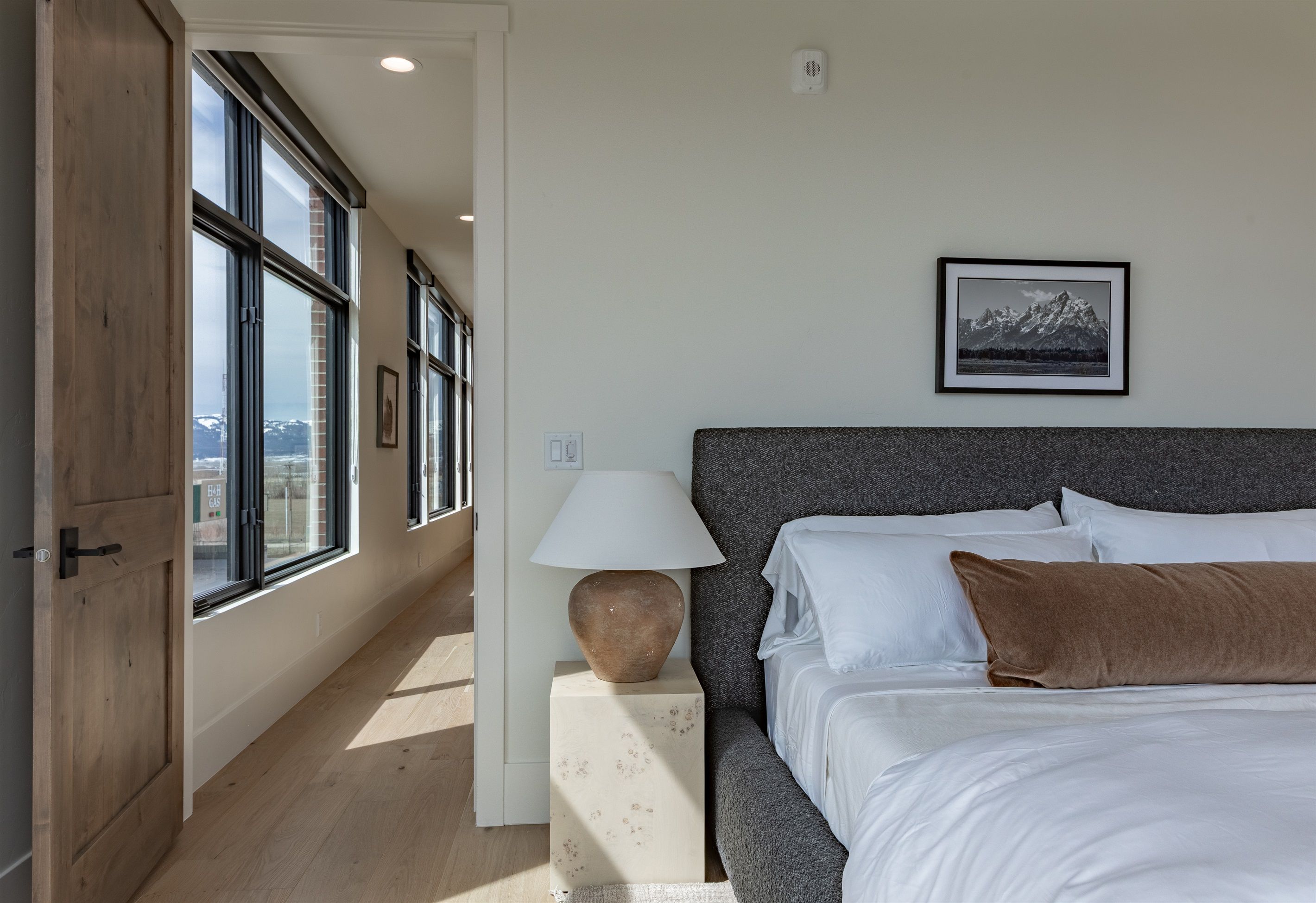 Un dormitorio con una cama grande, una mesita de noche, una lámpara, un cuadro en la pared y vistas al exterior.