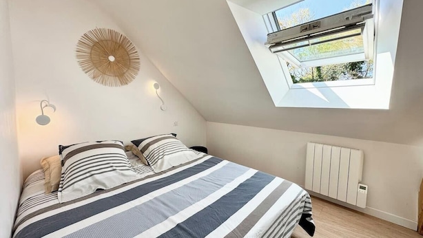 A bedroom with a sloped ceiling, a skylight, a bed with striped bedding, a wall-mounted heater, and a decorative wall piece.