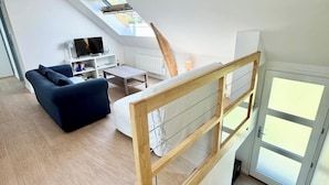 A modern loft apartment with a wooden staircase, a blue sofa, a dining table, and a TV in the living area.