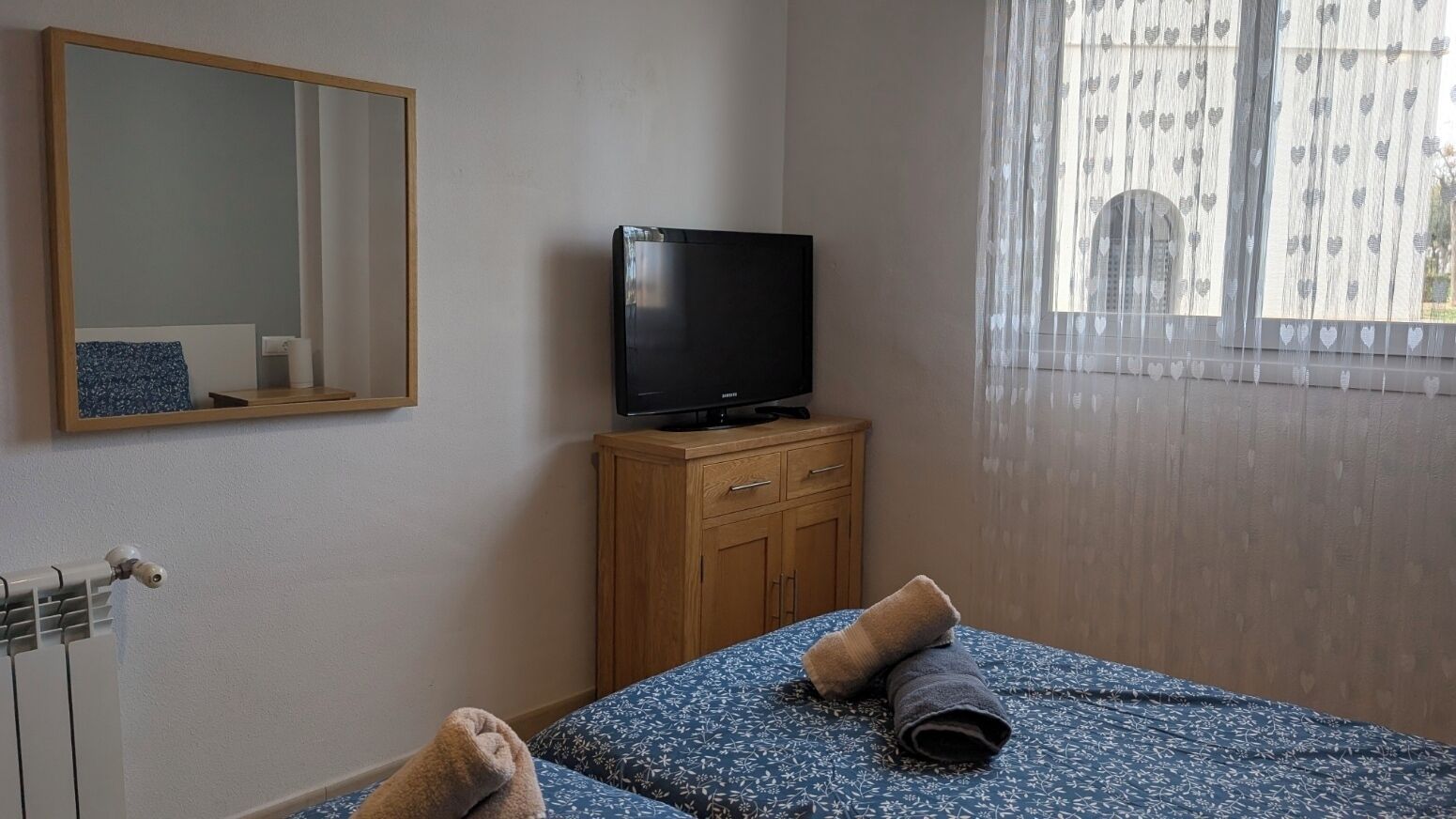 A bedroom with a bed, a television on a wooden cabinet, a mirror on the wall, and a window with heart-patterned curtains.