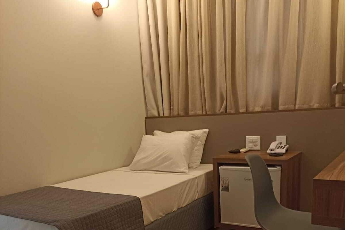 Una habitación de hotel con cama, escritorio con silla, mesita de noche y lámpara de pared.