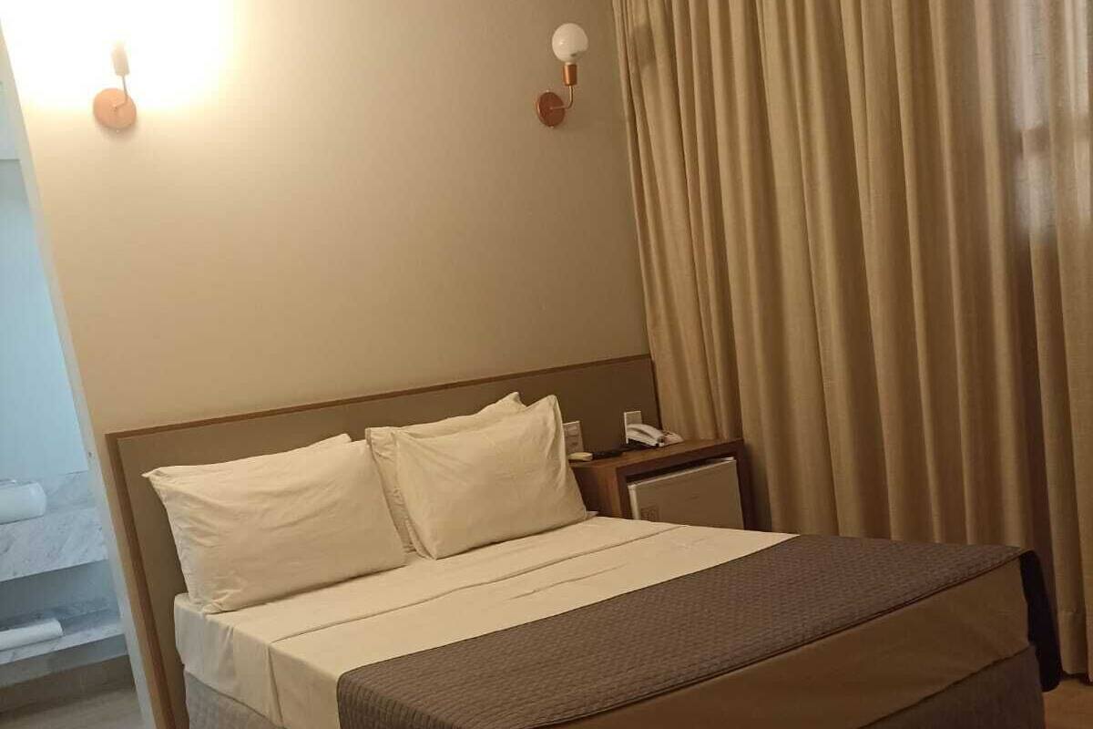 Una habitación de hotel con una cama, una mesita de noche, una lámpara y una cortina.