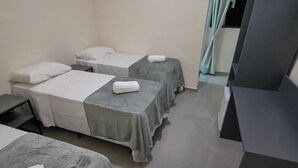 Une chambre d’hôtel avec trois lits simples, chacun recouvert d’une couverture grise et agrémenté d’oreillers blancs. Un petit bureau et une chaise se trouvent dans un coin, et un meuble de couleur sombre est adossé au mur.