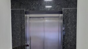 Un ascenseur moderne aux finitions métalliques, installé dans un couloir aux murs en granit foncé et au sol de couleur claire.