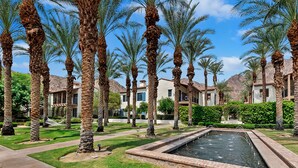 Un área ajardinada con palmeras, una piscina y un edificio con múltiples ventanas y balcones.
