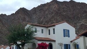Un edificio blanco con contraventanas azules y techo de tejas, rodeado de vegetación y montañas.