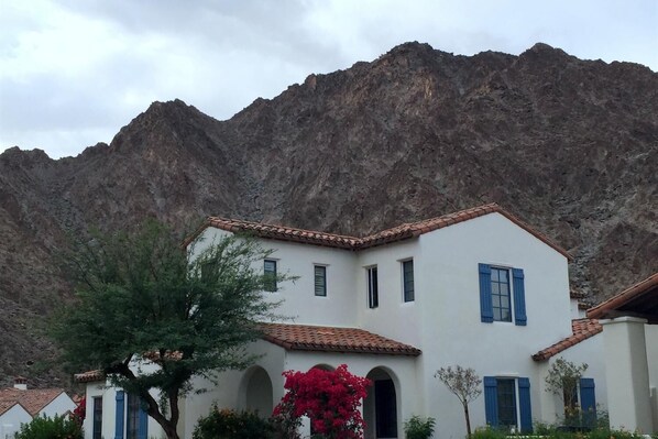 A white building with blue shutters and a tiled roof, surrounded by greenery and mountains.