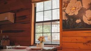 A wooden interior with a dining table set for two, a window with a floral painting, and a hanging light fixture. - White-Tail Nook Blue Ridge Cabin! (Blue Ridge)