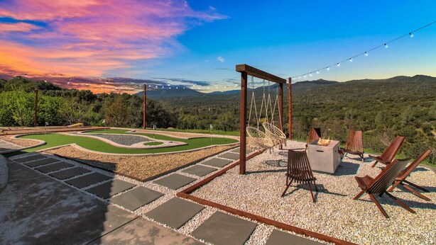 Eine Terrasse mit Feuerstelle, Sitzgelegenheiten und Blick auf einen angelegten Garten und Berge.