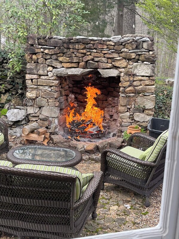 Una chimenea de piedra con fuego encendido, rodeada de muebles de mimbre y una mesa de vidrio.