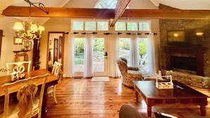 Een ruime woonkamer met een open haard, houten vloeren en een groot raam met uitzicht op een tuin.