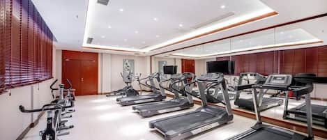 Academia com esteiras e bicicletas de exercício, um espelho grande e uma televisão na parede.