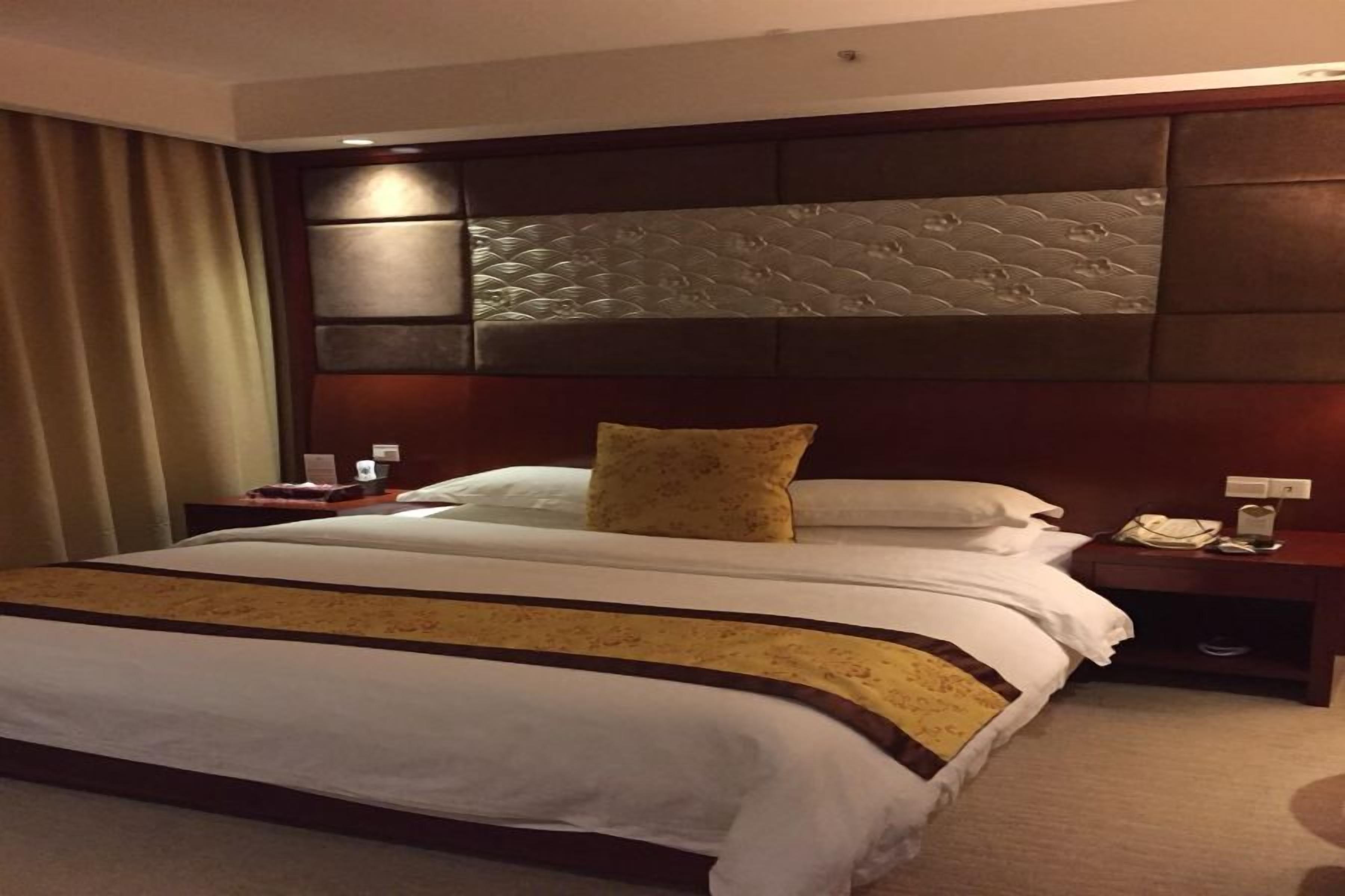 A hotel room with a large bed, a headboard with a textured design, a bedside table with a lamp, and a curtain on the left.