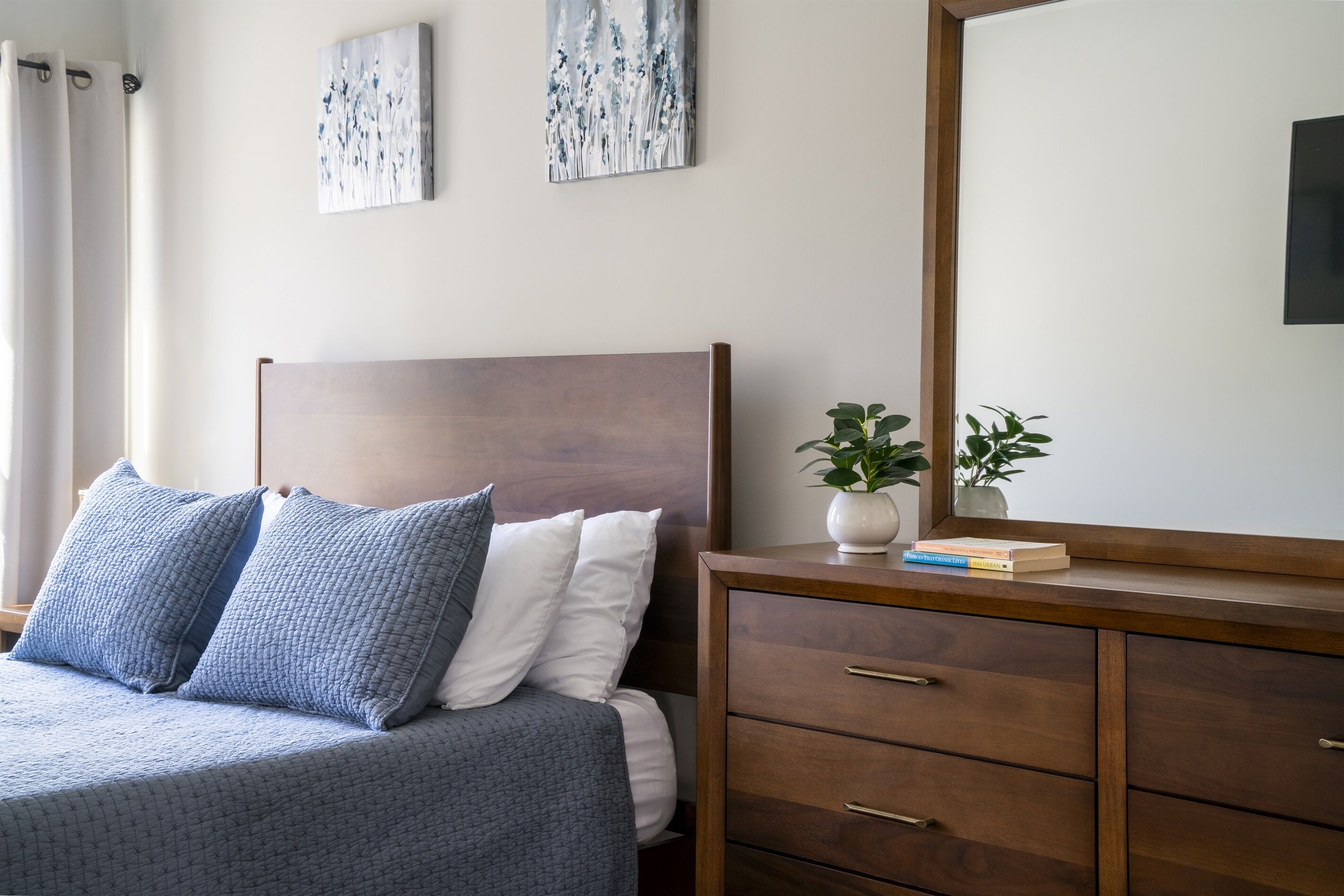 A bedroom with a bed, two pillows, a nightstand with a plant, and a mirror.