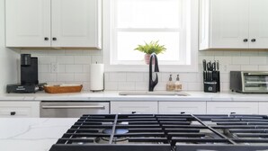 Una cocina moderna con armarios blancos, estufa a gas, cafetera, tostadora y una planta en maceta junto a la ventana.