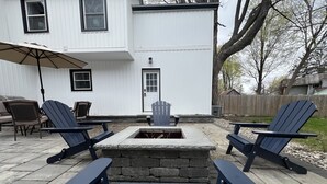 Un patio con sillas Adirondack, un hogar de piedra y una casa blanca al fondo.