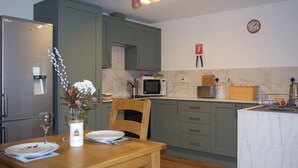 A kitchen with a dining table, chairs, a refrigerator, a microwave, a toaster, and a clock on the wall. - Beech Cottage - UK49564 (Sneaton, near Whitby)
