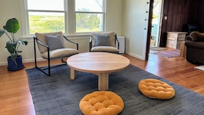 A living room with a round wooden table, two armchairs, a potted plant, and a view of the outdoors through large windows.
