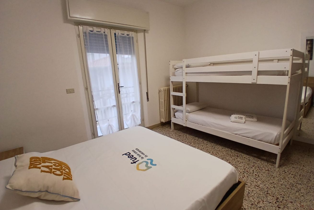 A bedroom with a bunk bed, a single bed with a pillow, a window with curtains, and a radiator.