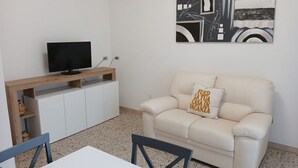 Een woonkamer met een beige bank, een televisie op een houten dressoir, een witte kast en een schilderij aan de muur.