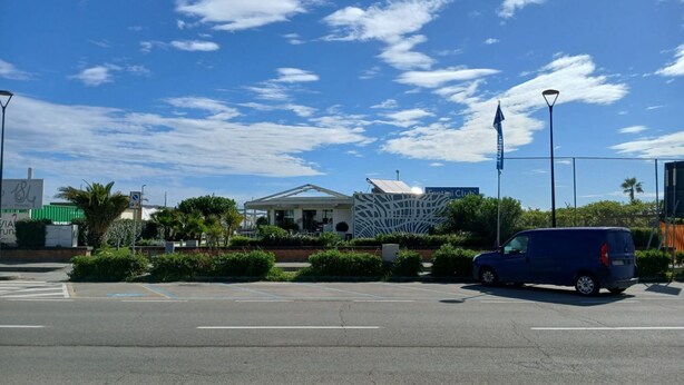 Een parkeerplaats met een blauwe bus, een gebouw met een decoratief gevelontwerp, en een heldere blauwe lucht met wolken.