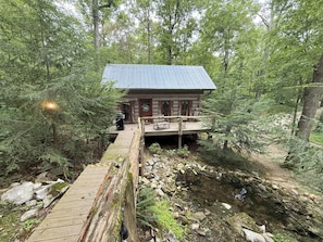 Une cabane dotée d’une terrasse, entourée d’une forêt dense et accessible par un pont en bois.