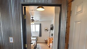 Een gang die leidt naar een slaapkamer met een bed, een plafondventilator en een raam met gordijnen.