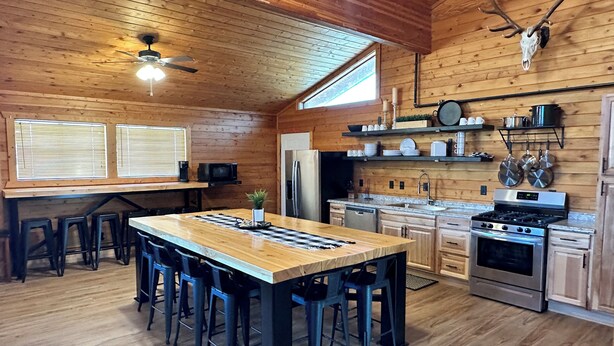 Een houten cabin met een eiland in het midden, een eettafel, barstoelen, een plafondventilator, een koelkast, een fornuis en een opgehangen hertenschedel.
