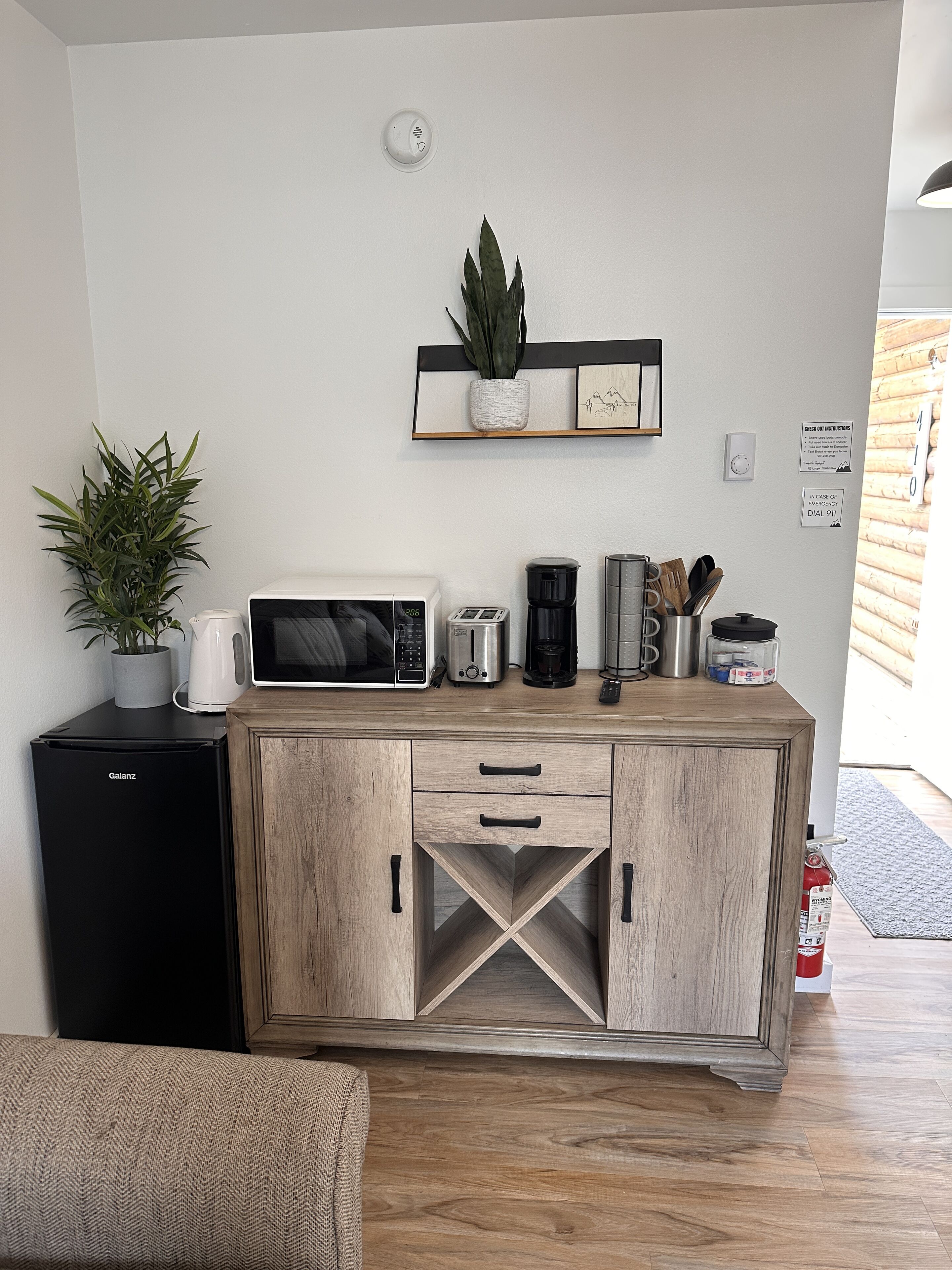 Una kitchenette compacta con microondas, tostadora, cafetera y un refrigerador pequeño. Hay un estante sobre la encimera con una planta y un cuadro enmarcado.