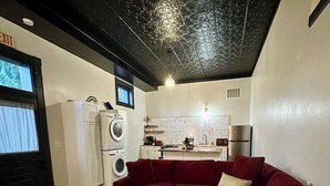 A compact living space with a red sofa, a small table, a washing machine, and a kitchenette. - Depot Suite - Ground-Floor Historic Loft on Katy Trail, 4 Mins to Hermann Wineries (Hermann)