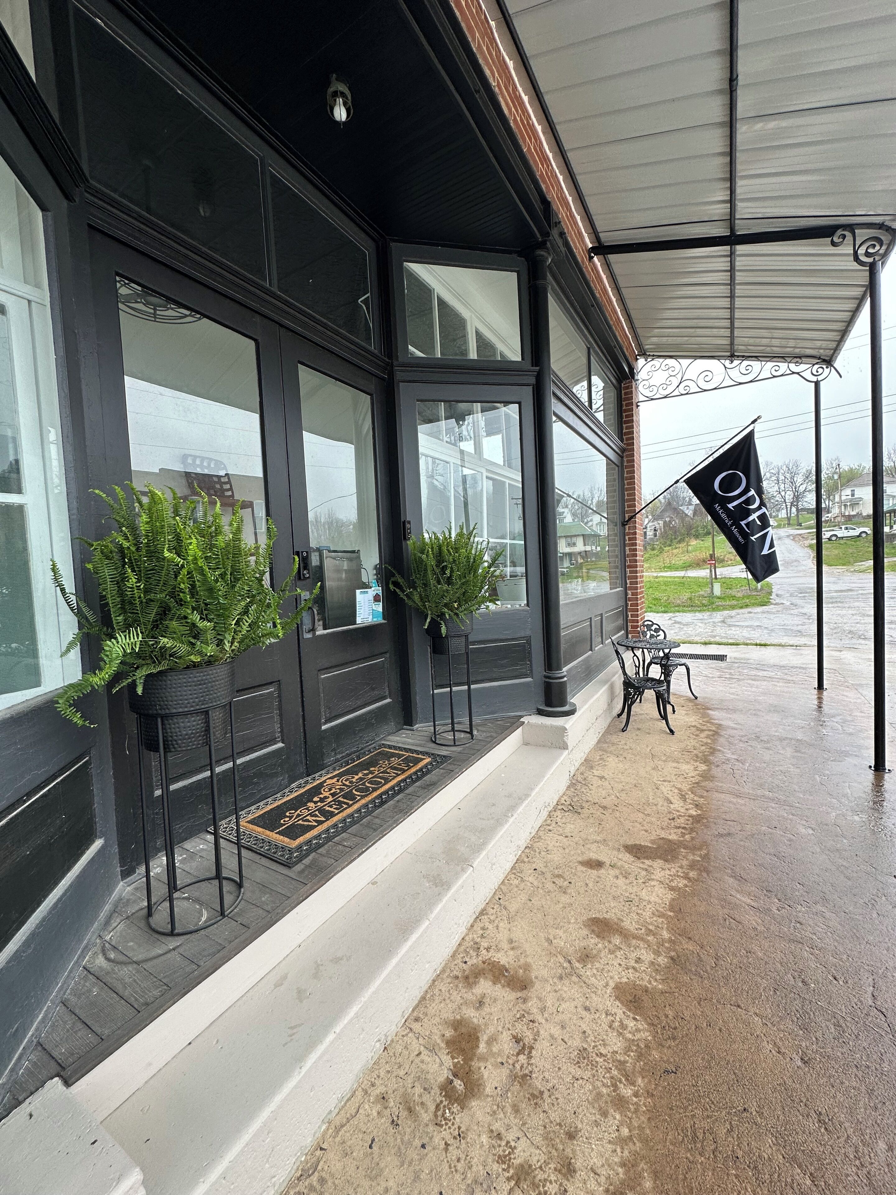 En sort bygning med store vinduer, en sort dør og en sort markise. Der er to krukkeplanter på trappen og et flag på markisen.