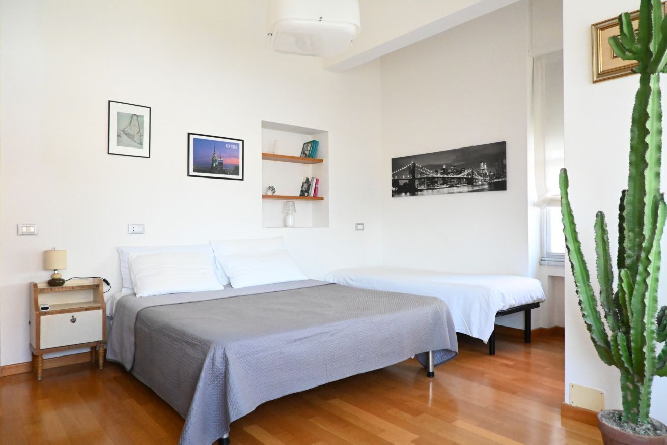 A bedroom with two beds, a nightstand, a shelf with books and a lamp, and a cactus plant.