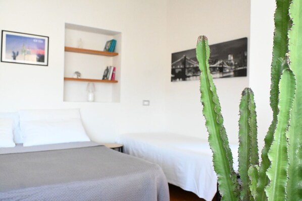 Un dormitorio con dos camas, una estantería con libros y un cuadro enmarcado, y un cactus en primer plano.