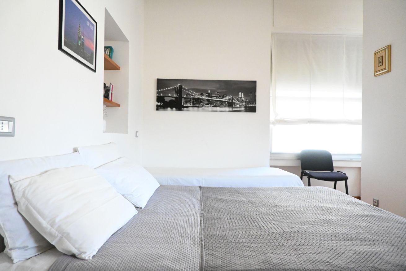 Een slaapkamer met een bed, een stoel, een raam met gordijnen, een aan de muur hangend schilderij en een plank met spullen.