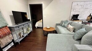 A modern living room with a grey sofa, a TV on a stand, a coffee maker, and a small wooden table. - Katy Suite - Chic 2BR Stay on Katy Trail in Historic Mercantile near Hermann (Hermann)